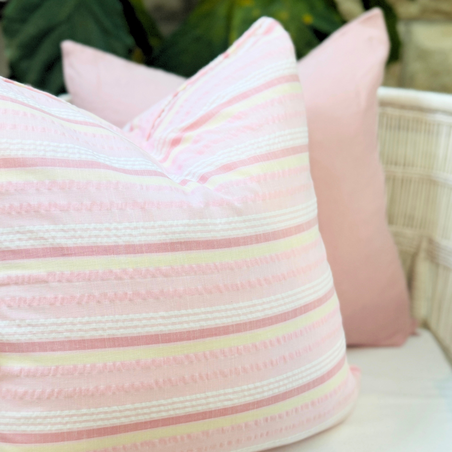 Pink striped cushion on a white surface with a blurred green plant in the background.