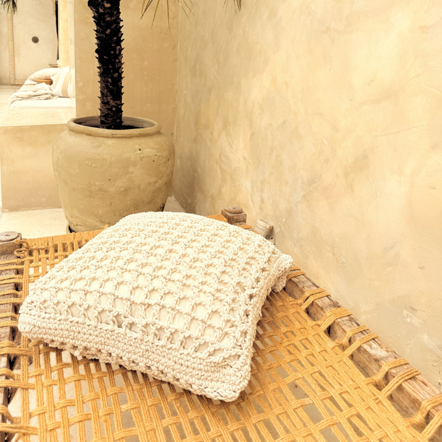 Woven chair with a textured cushion against a textured wall and potted plant.