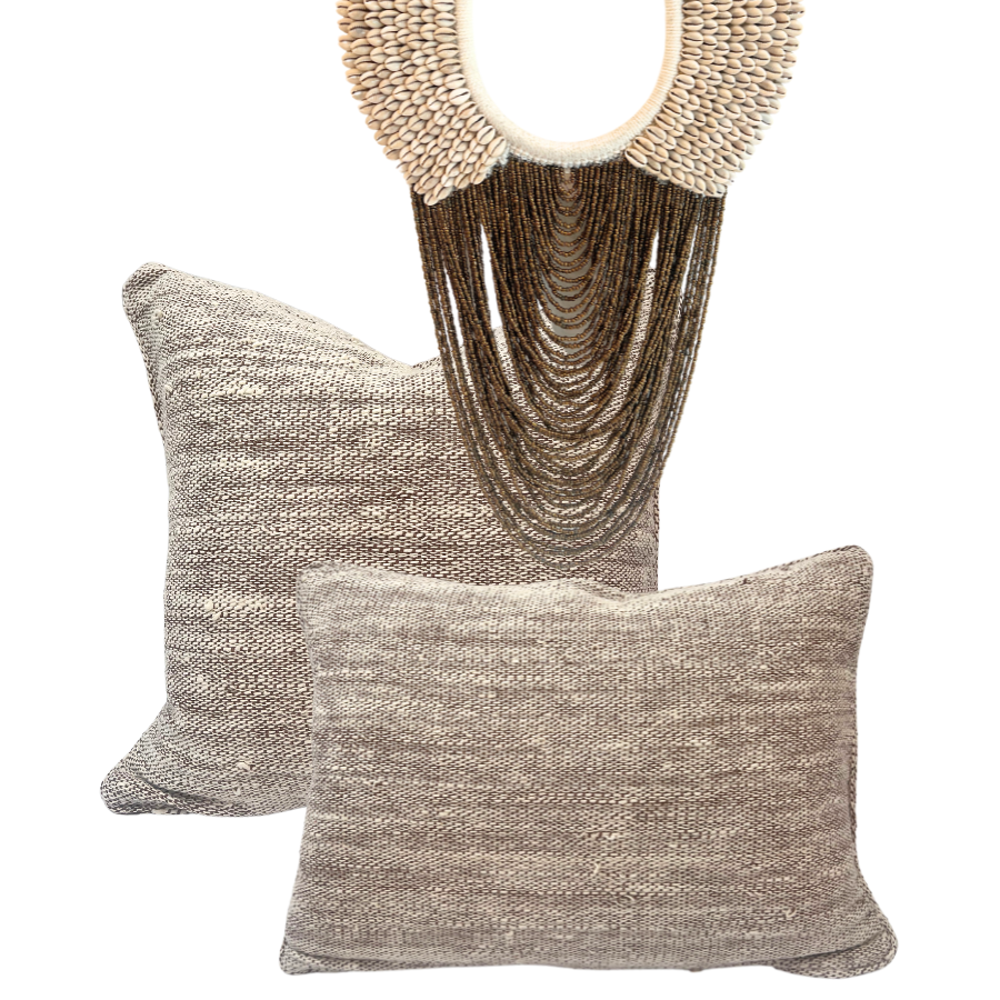 Two handwoven cushion covers in beige and brown tones with a large cowrie shell necklace wall hanging.