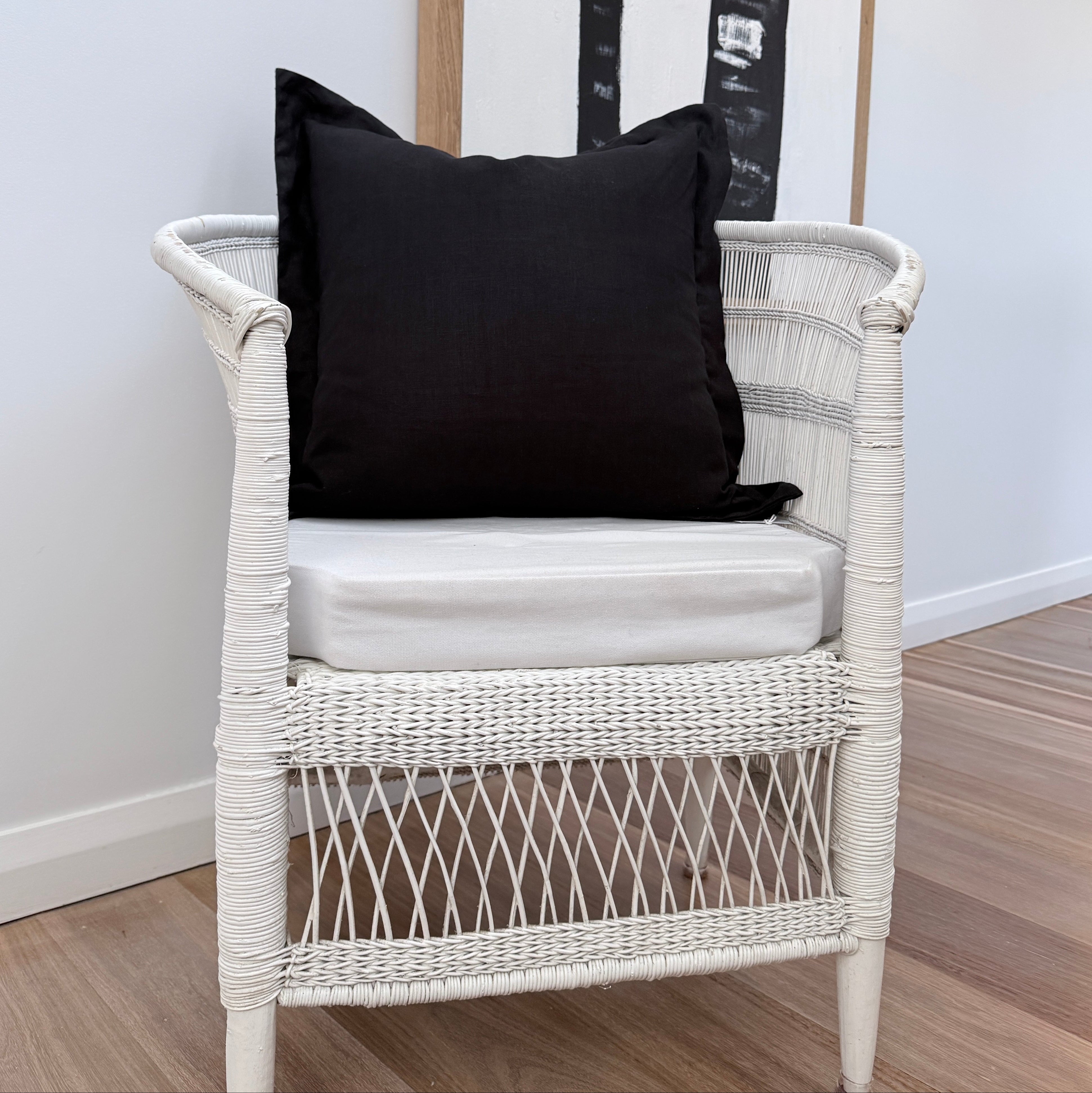 A white woven rattan armchair with a white cushion, styled with a black throw cushion, set against a white wall with a framed palm tree print.