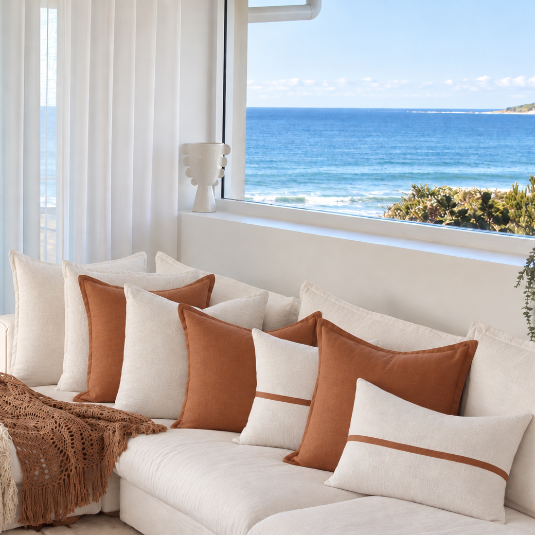 Cinnamon and cream linen cushions arranged on a white sofa in a coastal room overlooking ocean views.