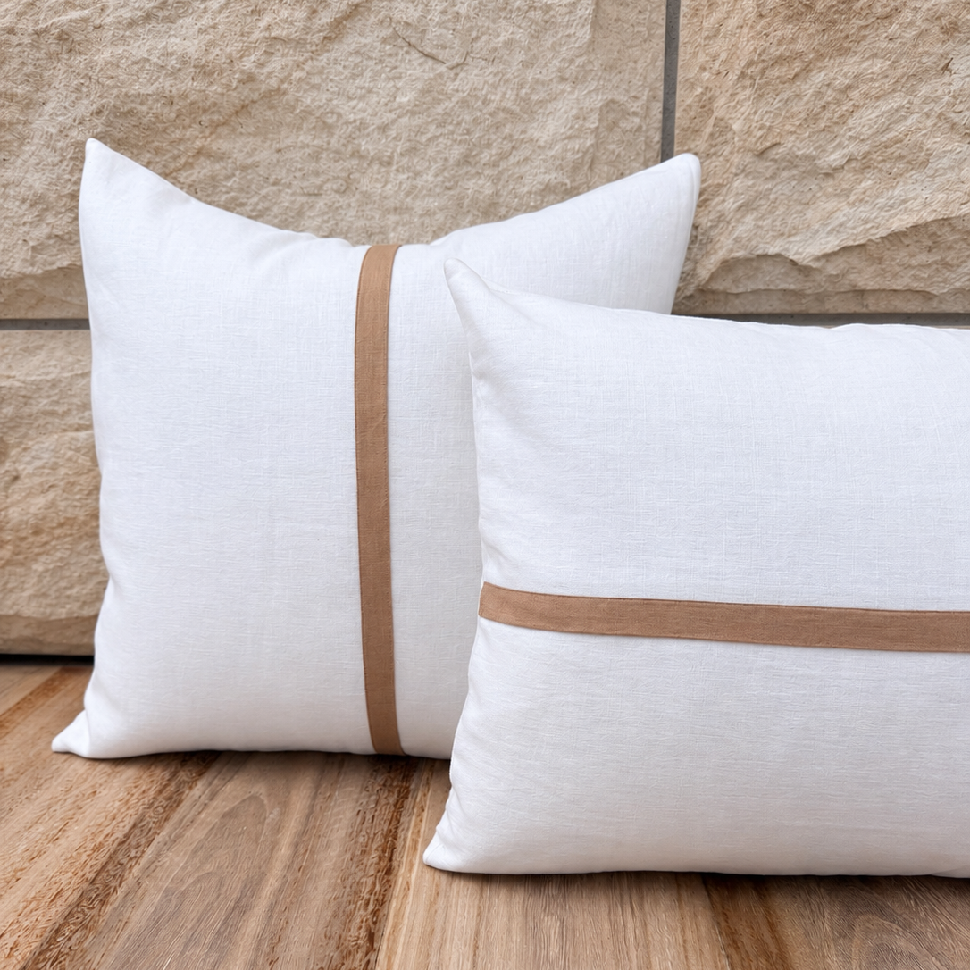 Two white linen throw pillows with horizontal tan stripes, one standing upright and one lying flat, set on a wooden floor against a stone wall.