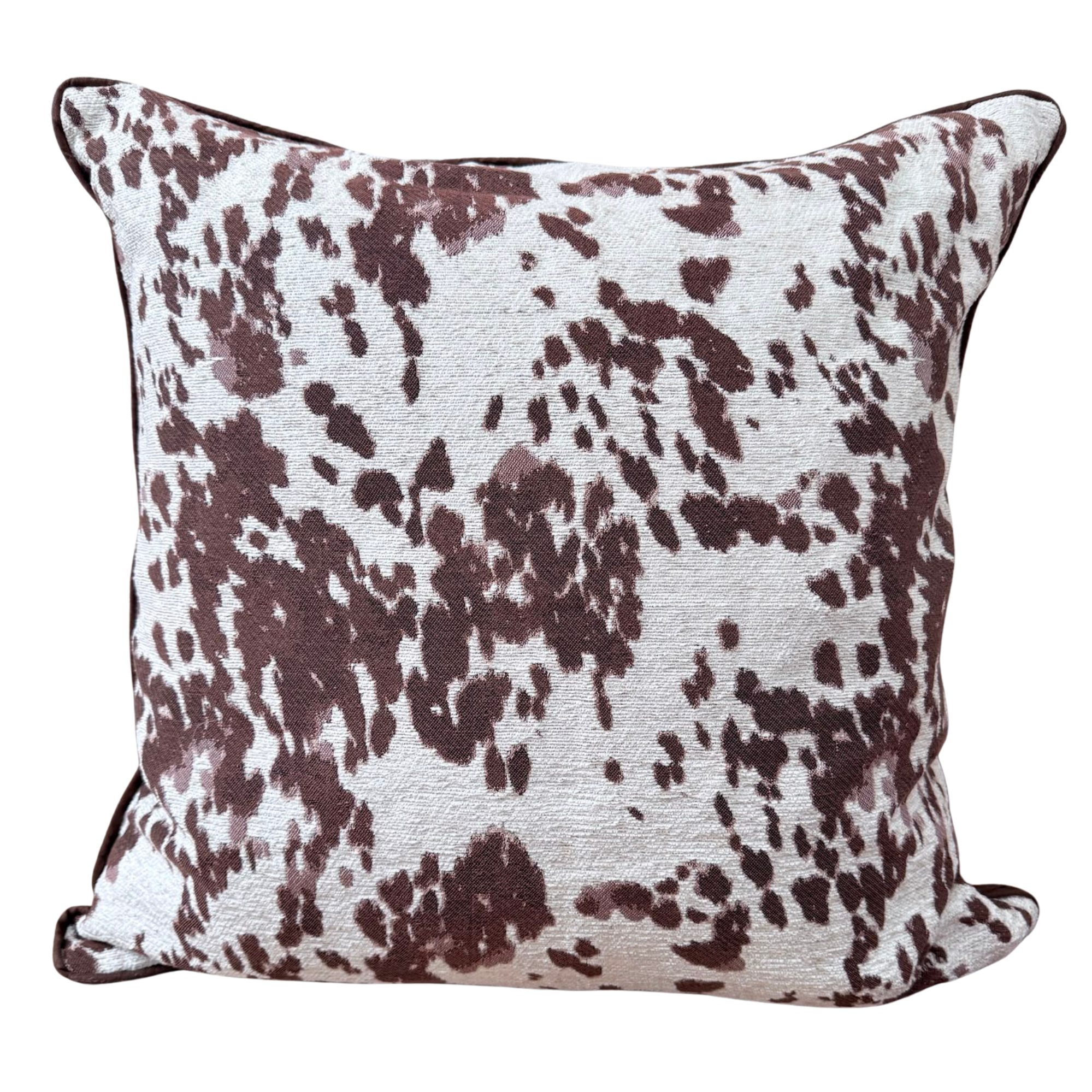 A square throw cushion featuring a brown and white cow-print pattern with dark brown piped edges, shown against a plain white background.