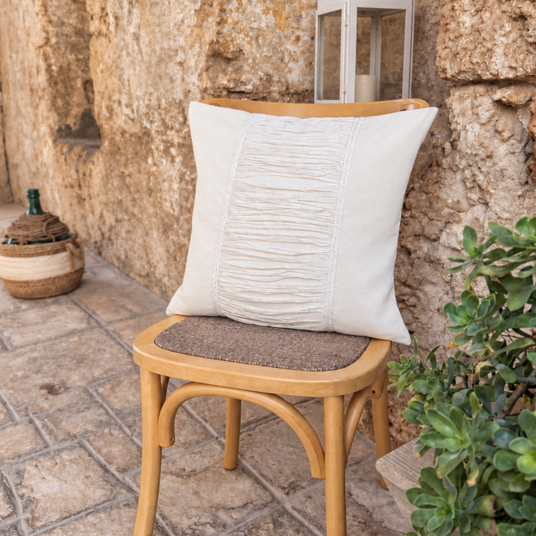 A square off-white throw pillow with vertical ruching on the front, displayed on a light wood bentwood chair in a rustic stone patio setting.