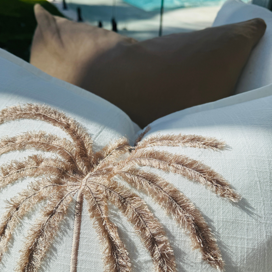 Decorative cushion with palm leaf design on a white surface, with a blurred background of a pool and chairs.