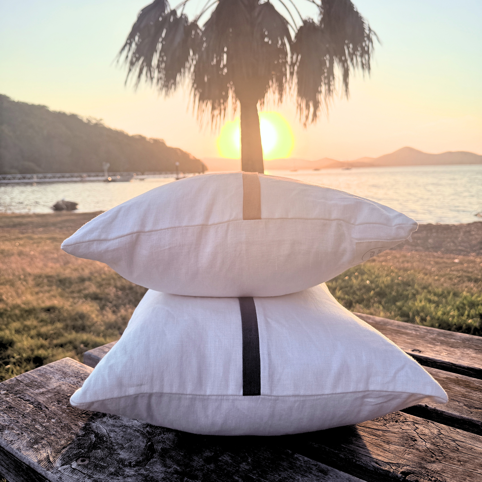 Two white cushion stacked on a wooden surface with a scenic sunset over water and palm tree in the background.